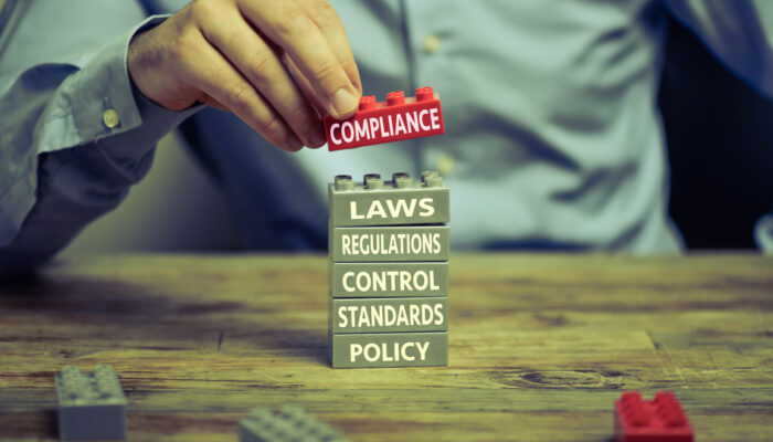 Man in business shirt playing with toy blocks on wood desk. There are compliance related words printed on the blocks